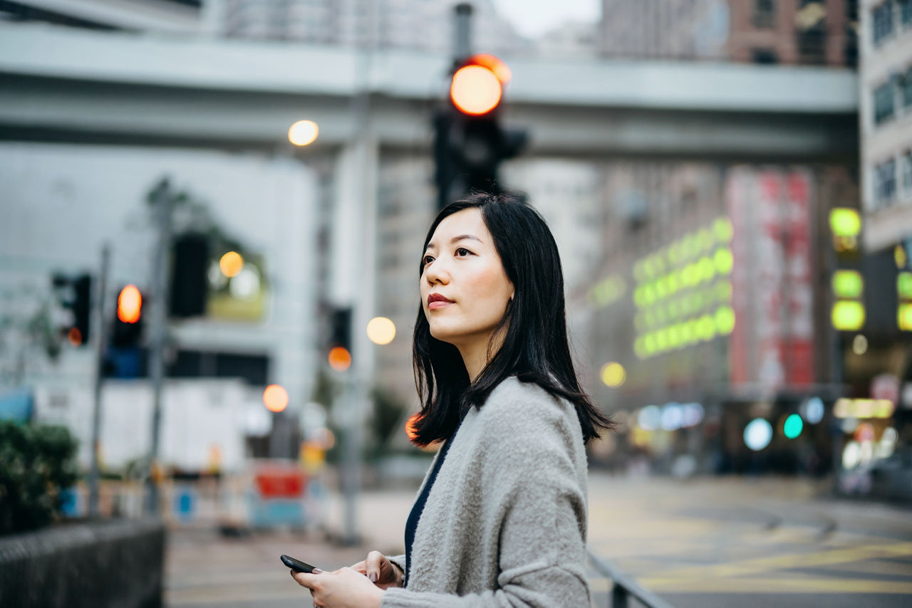 Young woman with smartphone commuting in the city, against busy city traffic and highrise buildings